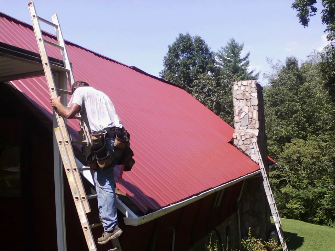 Roofer on a ladder beside a red metal roof and stone chimney — Soffit & Fascia Repair by Roofing Brothers in Orrstown